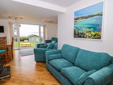 A living room with teal sofas a wooden dining table and chairs a large window overlooking a garden with a green shed at 3 Moranedd in Moelfre