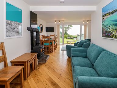 A living room with blue sofas a dining table wooden chairs a wood stove and sliding glass doors to a garden at 3 Moranedd in Moelfre