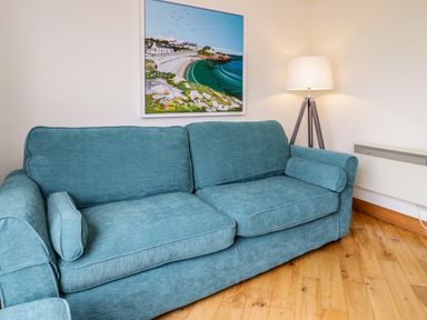 A blue sofa with cushions next to a floor lamp and a framed coastal painting on the wall at 3 Moranedd in Moelfre