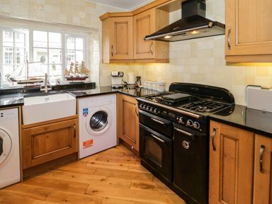 A kitchen with wooden cabinets a black stove a white washing machine a white sink and model ships on the windowsill at 3 Moranedd in Moelfre