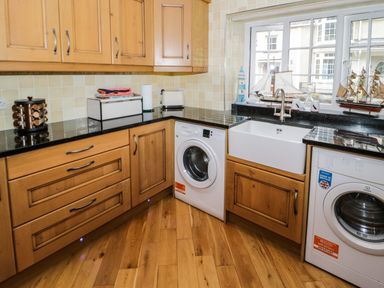 A kitchen with wooden cabinets black countertops two washing machines a white sink and model ships on the windowsill at 3 Moranedd in Moelfre