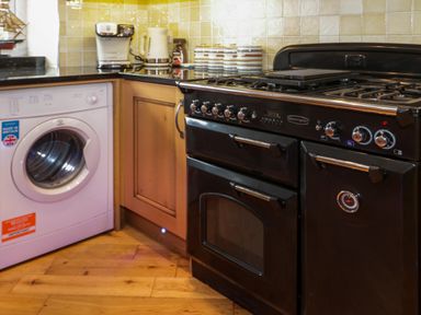 A kitchen corner with a washing machine a black stove wooden cabinets and countertop appliances at 3 Moranedd in Moelfre
