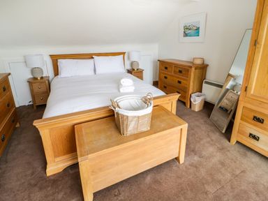 A bedroom with a double bed white linens wooden furniture bedside tables lamps a chest a wicker basket and a mirror at 3 Moranedd in Moelfre