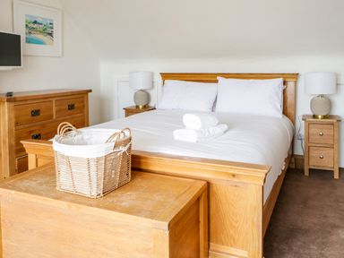 A bedroom with a wooden bed white bedding two bedside tables with lamps a wooden chest a basket on the chest and a dresser on the side at 3 Moranedd in Moelfre