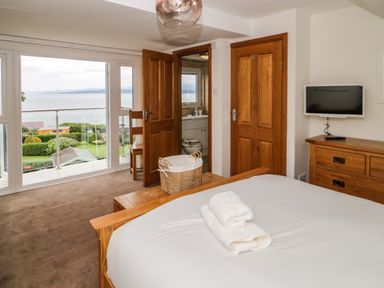 A bedroom with a wooden bed and dresser and a television mounted on the wall and an open door showing a bathroom and a large window with a view of the sea at 3 Moranedd Moelfre