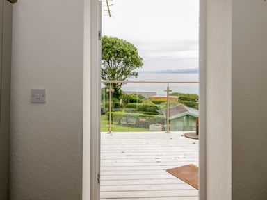 A view through a doorway onto a balcony with glass railing overlooking a garden and sea at 3 Moranedd in Moelfre