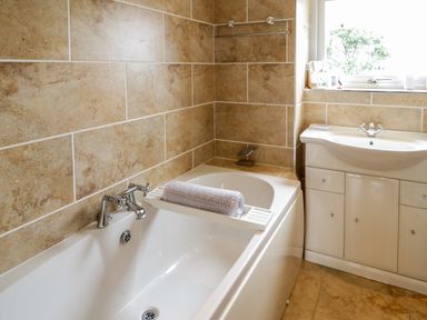 A bathroom with a bathtub a rolled towel on a bath tray a sink with storage cabinets and a window at 3 Moranedd Moelfre