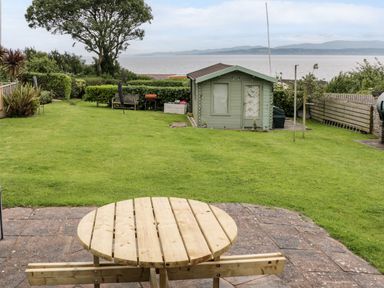 A garden with a wooden picnic table a shed hedge trees and a view of the sea at 3 Moranedd in Moelfre