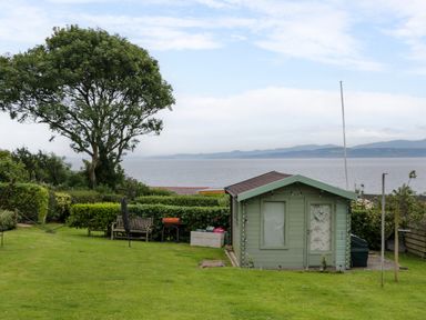 A garden with a small shed a wooden bench a barbecue and a tree overlooking the sea at 3 Moranedd in Moelfre