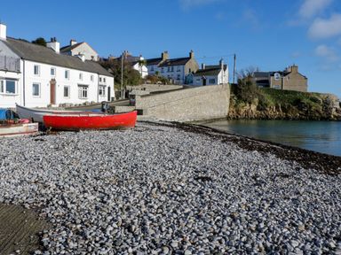 A pebble beach with red and white boats near white houses and a stone wall by the water at 3 Moranedd in Moelfre