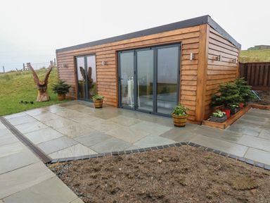 A wooden cabin with sliding glass doors on a stone patio with potted plants and a carved wooden owl statue outside at Height End Farm Log Cabin Rossendale near Bacup
