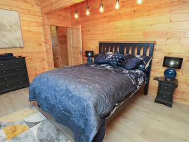 A bedroom with a bed with blue bedding two bedside tables with lamps a black dresser and wooden walls at Height End Farm Log Cabin in Rossendale near Bacup