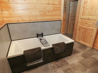 A bathroom with a rectangular bathtub with two towels hanging on it wooden paneled walls and a wooden door at Height End Farm Log Cabin in Rossendale near Bacup
