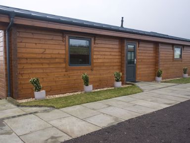 A wooden cabin exterior with potted plants along the wall and a paved walkway at Height End Farm Log Cabin in Rossendale near Bacup