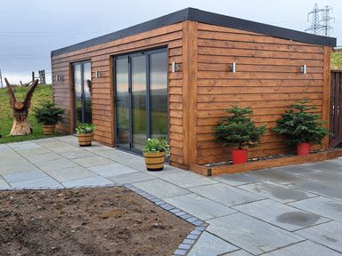 A wooden cabin with glass sliding doors surrounded by potted plants and a stone patio at Height End Farm Log Cabin Rossendale near Bacup