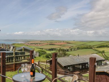 A patio table with a bottle and three glasses overlooking fields and a house at Three Views Lodge in Whitsand Bay Fort Holiday Village