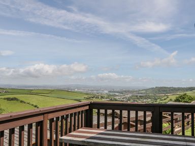 A wooden balcony railing and bench with a view of green fields and distant town at Three Views Lodge Whitsand Bay Fort Holiday Village