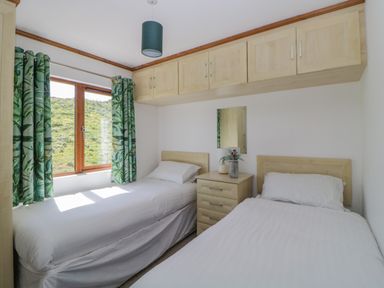 A bedroom with two single beds a window with green patterned curtains and overhead cabinets at Three Views Lodge in Whitsand Bay Fort Holiday Village