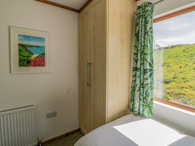 A bedroom corner with a wooden wardrobe next to a window with green leafy curtains and a bed below the window at Three Views Lodge in Whitsand Bay Fort Holiday Village