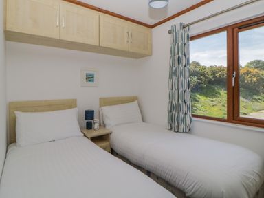 A bedroom with two single beds side by side a small bedside table a window with curtains and overhead cabinets at Three Views Lodge in Whitsand Bay Fort Holiday Village