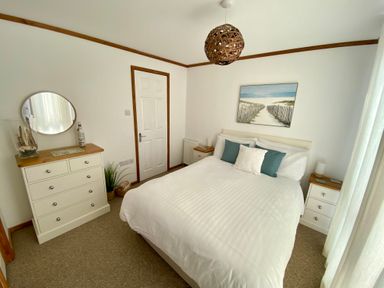 A bedroom with a double bed white bedding bedside tables a dresser with a round mirror and a painting above the bed at Three Views Lodge in Whitsand Bay Fort Holiday Village