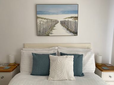 A bed with white and blue pillows and a beach painting above with bedside tables and lamps at Three Views Lodge in Whitsand Bay Fort Holiday Village