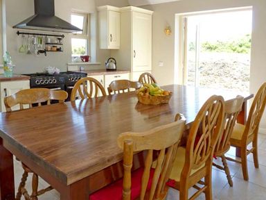 A kitchen with a wooden dining table and chairs a fruit bowl on the table and a black stove with cabinets at Bluebell Cottage in Spanish Point County Clare
