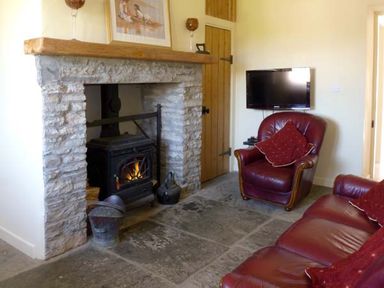 A living room with a stone fireplace wood burning stove red leather armchair and sofa and a wall mounted television at Bluebell Cottage in Spanish Point County Clare