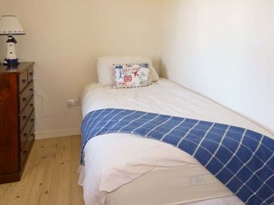 A bedroom with a single bed with white bedding and a blue checkered throw next to a wooden chest of drawers with a lighthouse lamp at Bluebell Cottage in Spanish Point County Clare
