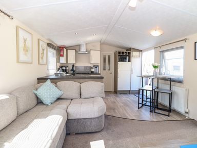 A living room with a beige sectional sofa a small dining table with two stools and a kitchen area at Hazell Thorne Hideaway in Fritton