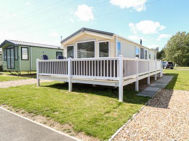 A beige mobile home with a white railing deck on a grassy plot with a gravel driveway at Hazell Thorne Hideaway in Fritton