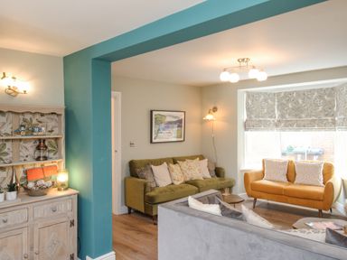 A living room with green and orange sofas a decorative cabinet and floral window blinds at Bay Cottage in Weymouth