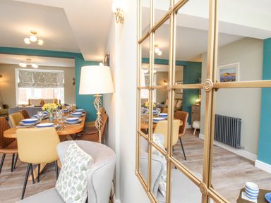 A dining area with a wooden table set with plates and glasses surrounded by chairs and a living room visible through an open space at Bay Cottage in Weymouth