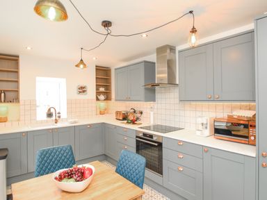 A kitchen with grey cabinets a wooden table blue chairs and various appliances at Bay Cottage in Weymouth
