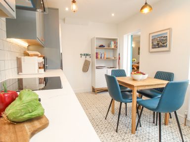 A kitchen with a wooden dining table and blue chairs next to shelves with books and a framed picture on the wall at Bay Cottage in Weymouth