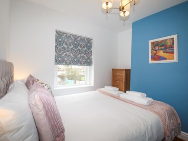 A bedroom with a bed, pillows, towels on the bed, a wooden dresser, a blue accent wall with a painting, and a window with patterned curtains at Bay Cottage in Weymouth