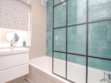 A bathroom with a bathtub and blue tiled wall glass shower screen and a sink with a floral window blind at Bay Cottage in Weymouth