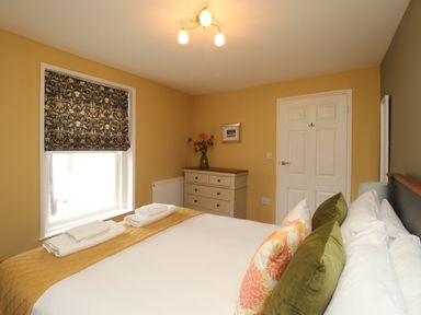 A bedroom with a bed with pillows and folded towels next to a window with patterned blind and a dresser with a vase of flowers at Bay Cottage in Weymouth