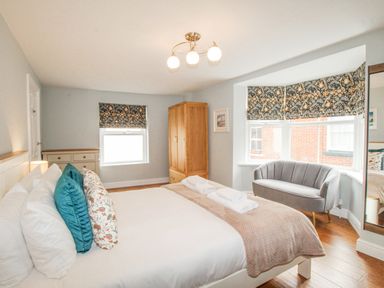 A bedroom with a bed with cushions and folded towels a grey sofa wooden wardrobe and windows with patterned blinds at Bay Cottage in Weymouth