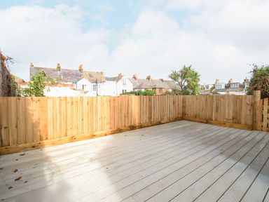 An empty wooden deck area surrounded by a wooden fence with rooftops and trees in the background at Bay Cottage in Weymouth