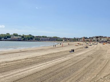 A sandy beach with people sitting and walking along the shore near buildings in Weymouth