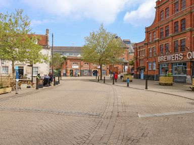 A cobblestone street with trees benches and people near red brick buildings at Brewers Quay in Weymouth