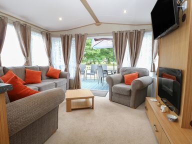 A living room with gray sofas and orange cushions a wooden coffee table a wall mounted television and an outdoor seating area at Stanleys Lodge Foxburrow Hang in Belton Norfolk