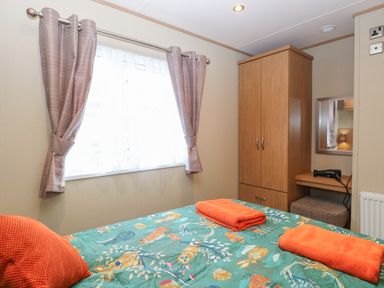 A bedroom with a bed covered in a green bird-patterned bedspread and orange towels and pillow next to a wooden wardrobe and a small vanity with a hairdryer at Stanleys Lodge Foxburrow Hang in Belton Norfolk