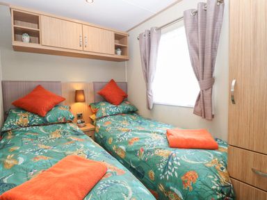 A bedroom with two single beds with green patterned bedspreads orange pillows and towels a wooden cabinet and shelving unit with plants and a window with curtains at Stanley's Lodge  Foxburrow Hang in Belton Norfolk