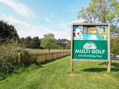 A green outdoor area with a wooden fence and a sign for multi golf and swimming pool at Stanleys Lodge Foxburrow Hang in Belton Norfolk