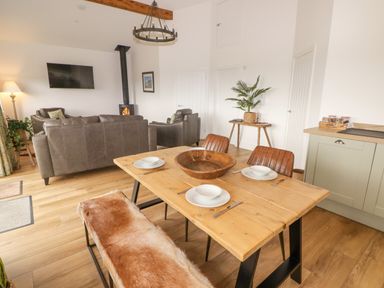 A dining area with a wooden table set with white dishes and a bench with a fur cover next to a living room with gray sofas a wall-mounted television and a wood-burning stove at Heritage Retreat in Monknash