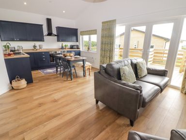 An open plan kitchen and living area with navy cabinets a wooden dining table with chairs a brown leather sofa large windows and wooden flooring at Monks Rest in Monknash