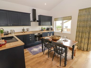 A kitchen with dark cabinets a wooden dining table with chairs and a window with patterned curtains at Monks Rest in Monknash