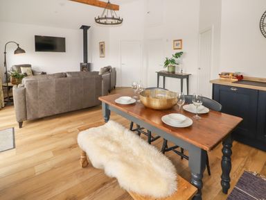 A dining area with a wooden table set for four and a bench with a fur cover next to a living room with leather sofas a wall-mounted television and a fireplace at Monks Rest in Monknash